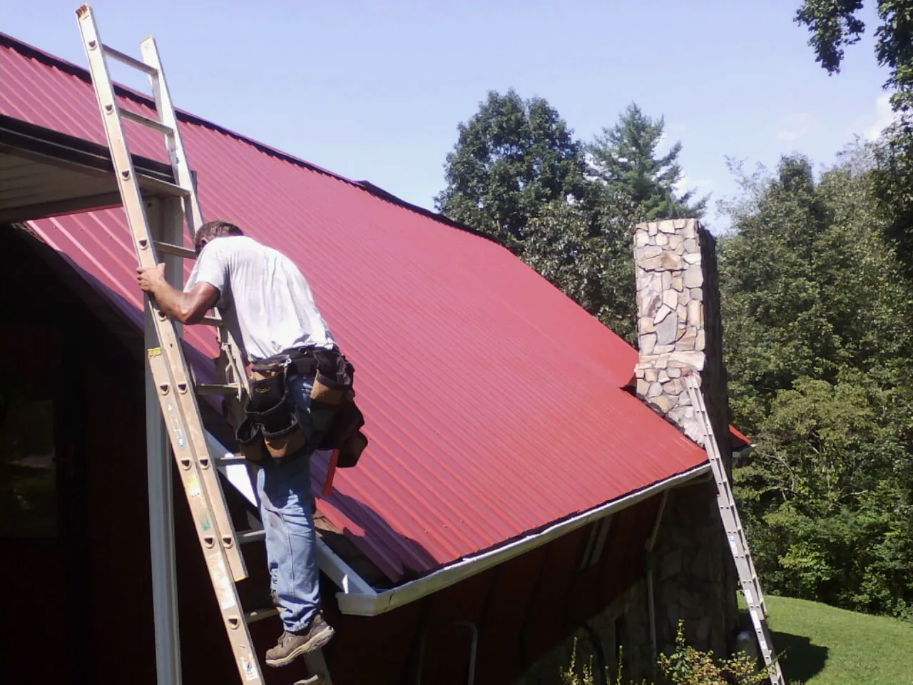 Roofer on a ladder beside a red metal roof and stone chimney — Soffit & Fascia Repair by Roofing Brothers in Sagaponack